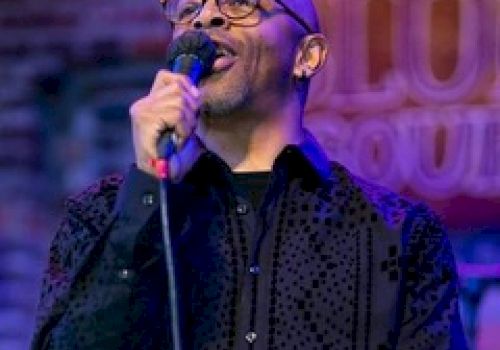 A person with glasses is holding a microphone, performing on stage with a sign in the background suggesting a jazz club or live music venue.