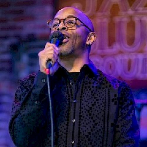 A person with glasses is holding a microphone, performing on stage with a sign in the background suggesting a jazz club or live music venue.